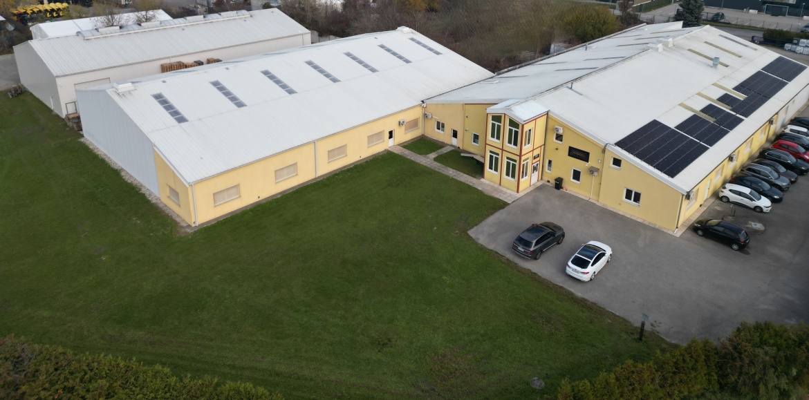 Foto aus der Vogelperspektive eines Industriebetriebs mit gelber Fassade, Parkpl&auml;tzen, Photovoltaikanlage auf dem Dach und einer gro&szlig;en Rasenfl&auml;che davor.